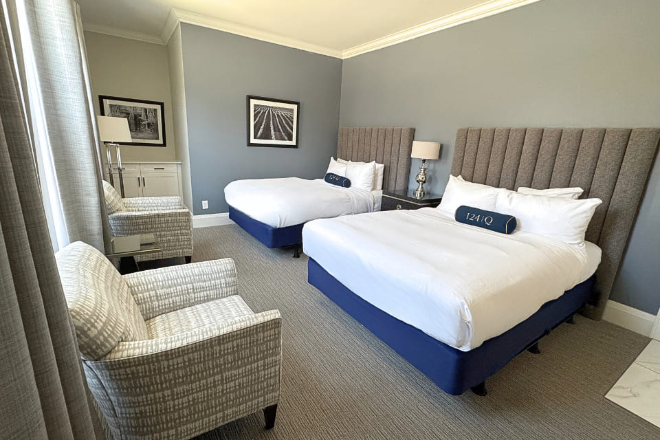 Hotel room with two double beds, gray upholstered headboards, white bedding, a nightstand with a lamp, two patterned armchairs, gray carpet, and framed photos on pale gray walls. Large windows bring in natural light. 124 on Queen Hotel and Spa | Hotel & Spa Niagara-on-the-lake | 1 (855) 988-4552