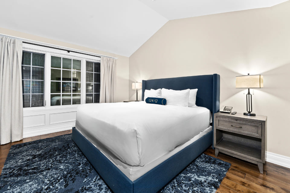 Modern bedroom with a blue upholstered bed, white bedding, two bedside tables with lamps, a large window with beige curtains, and a dark blue patterned rug on hardwood floors. 124 on Queen Hotel and Spa | Hotel & Spa Niagara-on-the-lake | 1 (855) 988-4552