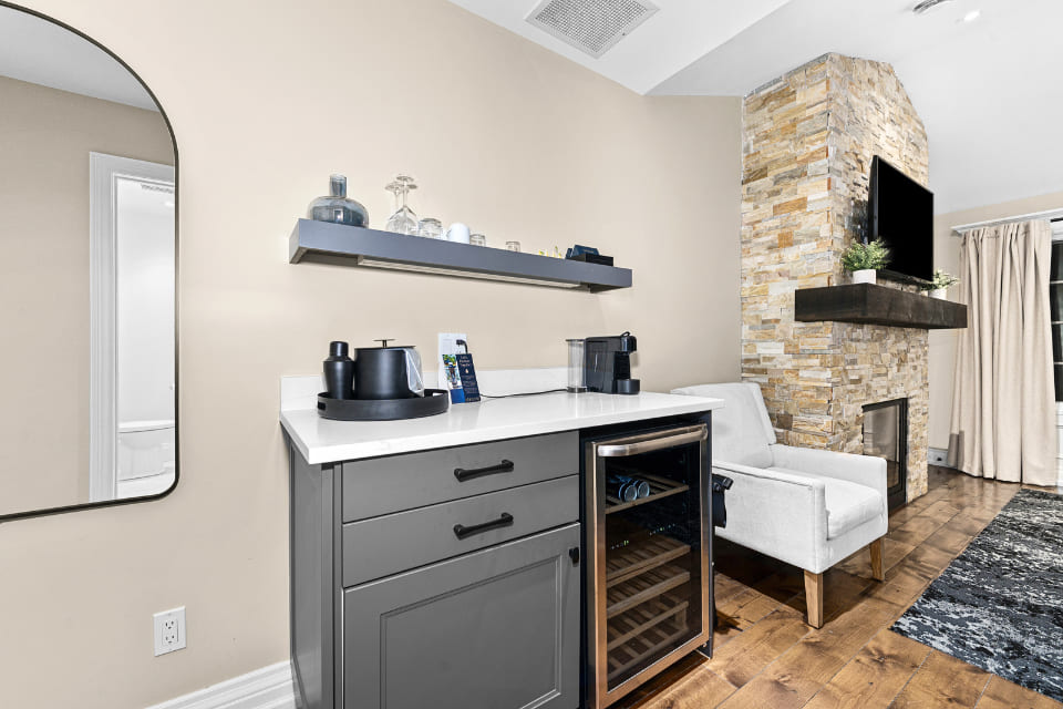 A modern kitchenette with gray cabinetry, a small wine fridge, a coffee maker, and glassware on a floating shelf; adjacent to a cozy seating area with a stone fireplace and armchair. 124 on Queen Hotel and Spa | Hotel & Spa Niagara-on-the-lake | 1 (855) 988-4552