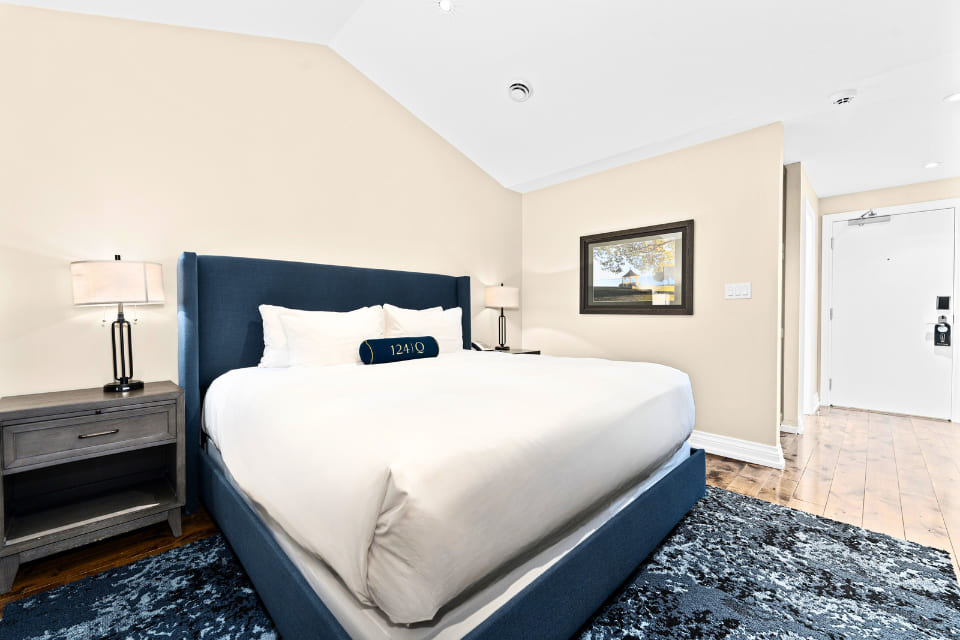 A modern hotel room with a large blue-upholstered bed, white bedding, two bedside tables with lamps, a framed landscape picture on the wall, and a patterned blue rug on a wooden floor. 124 on Queen Hotel and Spa | Hotel & Spa Niagara-on-the-lake | 1 (855) 988-4552