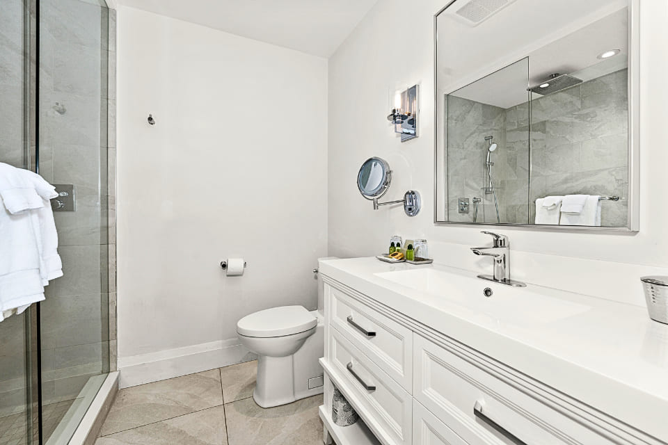 Modern bathroom with a glass shower, white toilet, and double-sink vanity with drawers. Large mirror above the sinks, wall-mounted light fixtures, and neutral tile floor create a clean, bright space. 124 on Queen Hotel and Spa | Hotel & Spa Niagara-on-the-lake | 1 (855) 988-4552